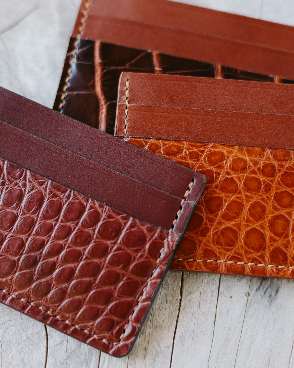 Three slim leather card holders with textured alligator belly and smooth leather panels in Bordeaux, cognac, and dark brown, arranged on a light wood surface.