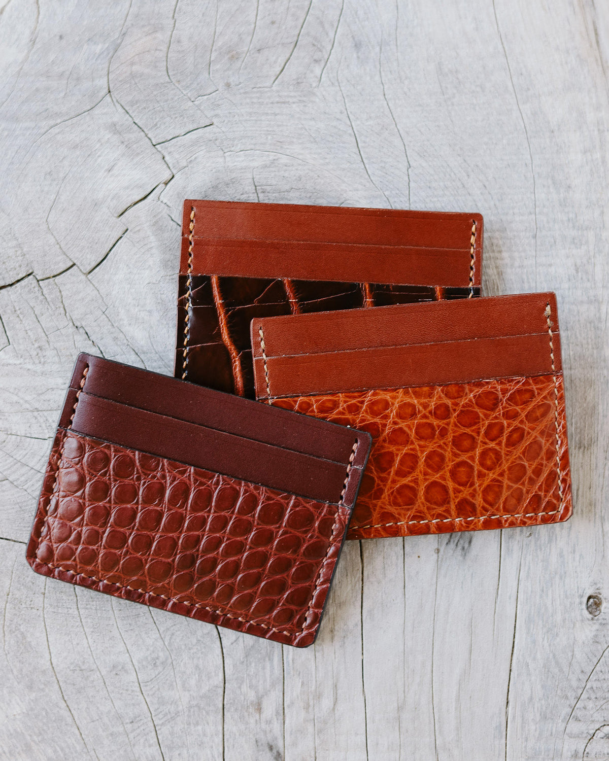 Three slim leather card holders in different brown tones and textures, arranged on a light gray wood surface.